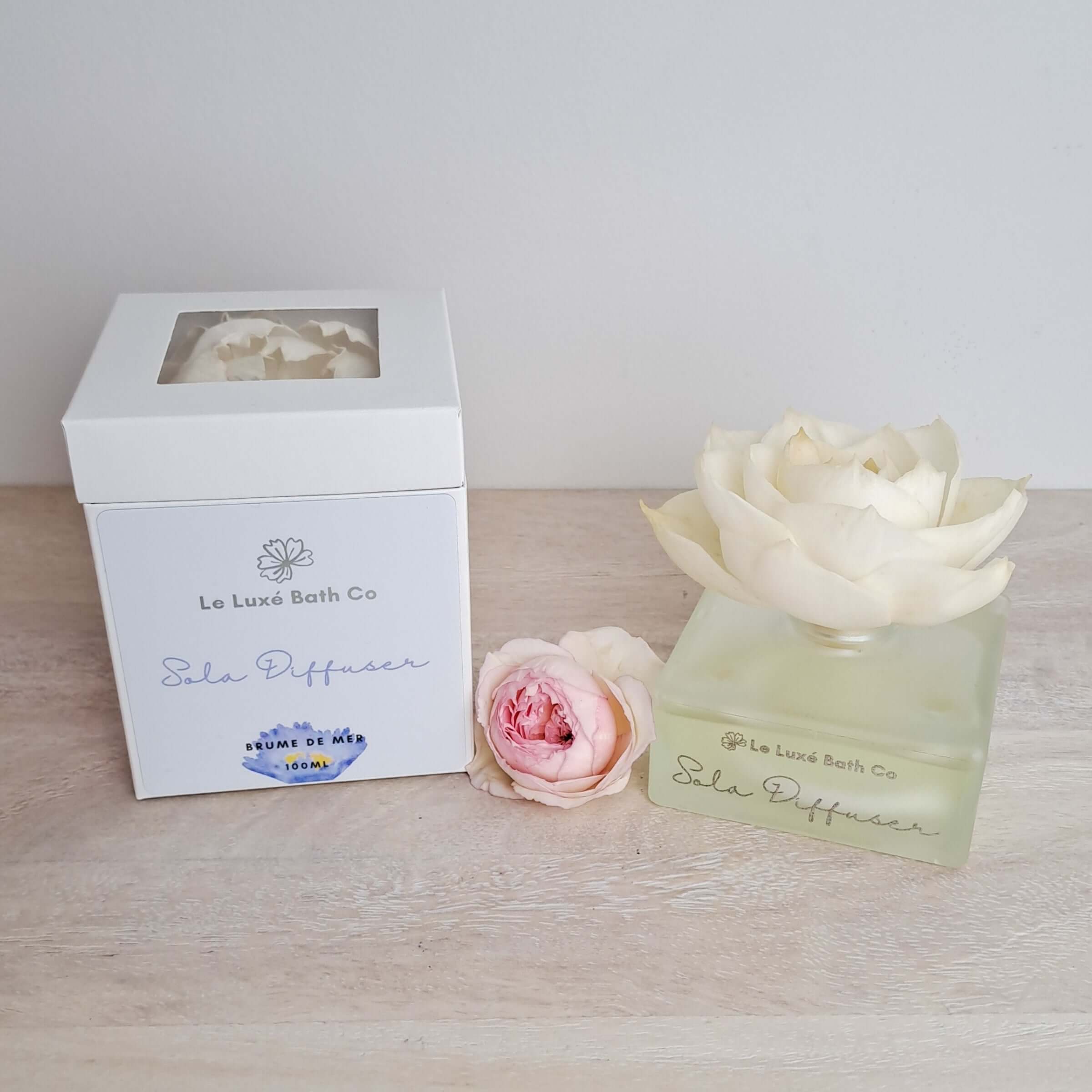 Le Luxe Bath Co Sola Fragrance Diffuser with packaging and flowers on a wooden surface