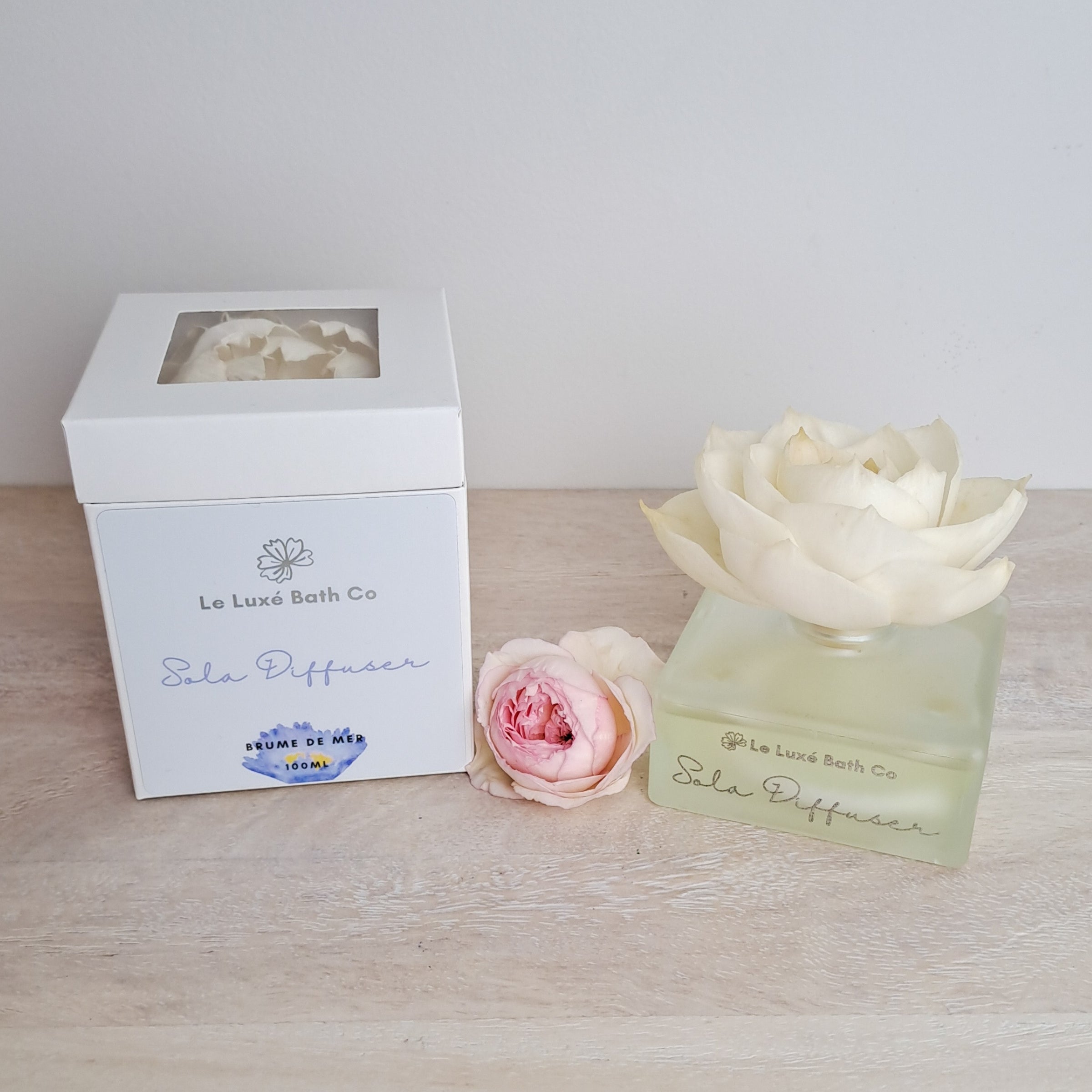 Le Luxe Bath Co Sola Fragrance Diffuser with packaging and flowers on a wooden surface